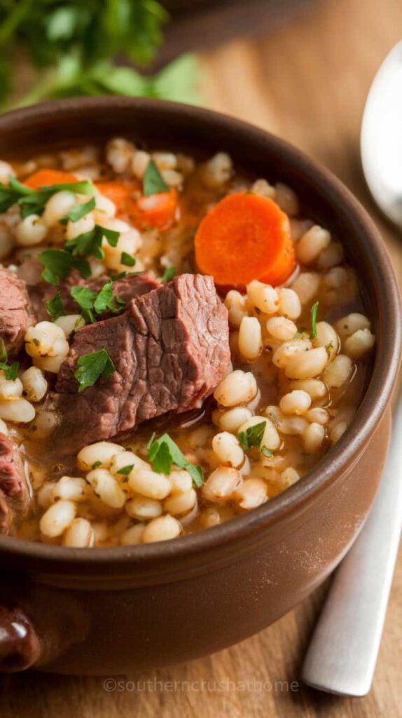 A bowl of beef barley soup with chunks of beef, barley, and carrots garnished with parsley.